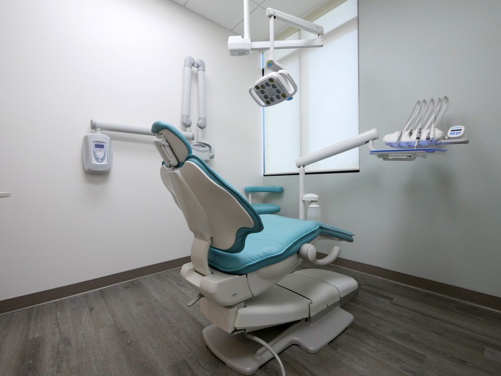 Photo of a modern dental treatment room with a blue and white dental chair, overhead lighting, and equipment.