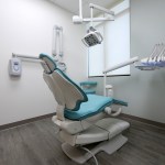 Photo of a modern dental treatment room with a blue and white dental chair, overhead lighting, and equipment.