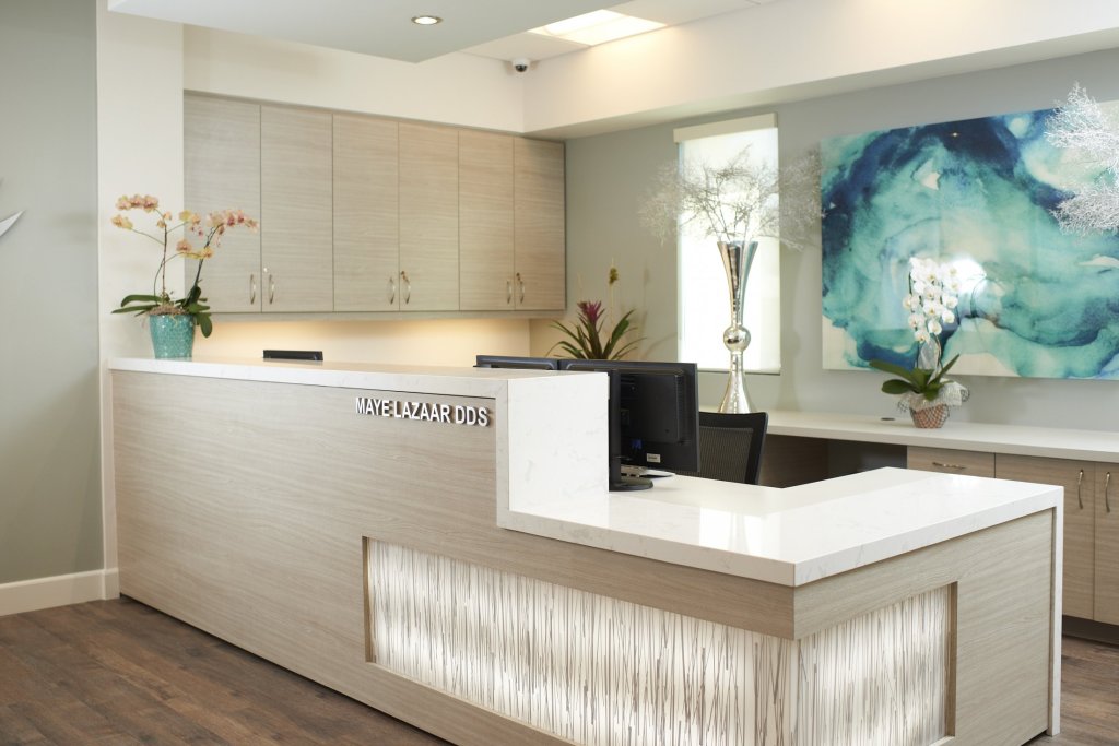 Photo of a clean, modern dental reception desk with white countertops, decorative plants, and a large wall art piece.