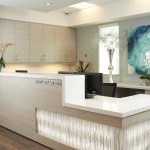 Photo of a clean, modern dental reception desk with white countertops, decorative plants, and a large wall art piece.
