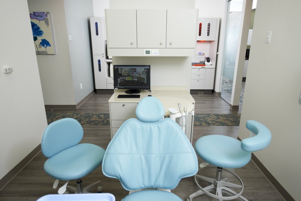 Photo of a dental treatment room with a blue chair, two side chairs, and medical equipment in a clean, professional setting.
