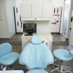 Photo of a dental treatment room with a blue chair, two side chairs, and medical equipment in a clean, professional setting.
